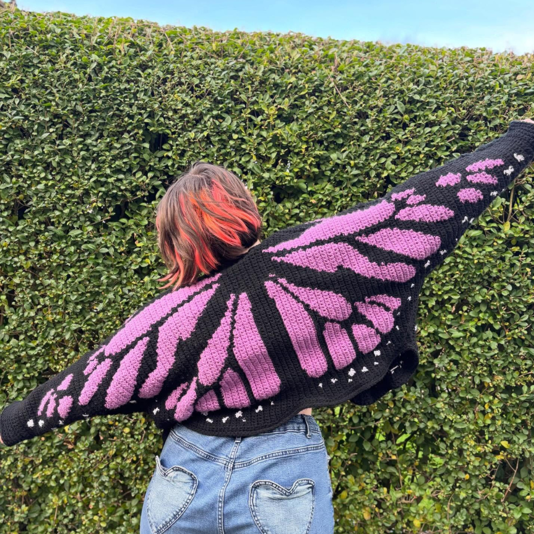 Person wearing a black crocheted cardigan with pink monarch butterfly wings against a green hedge.