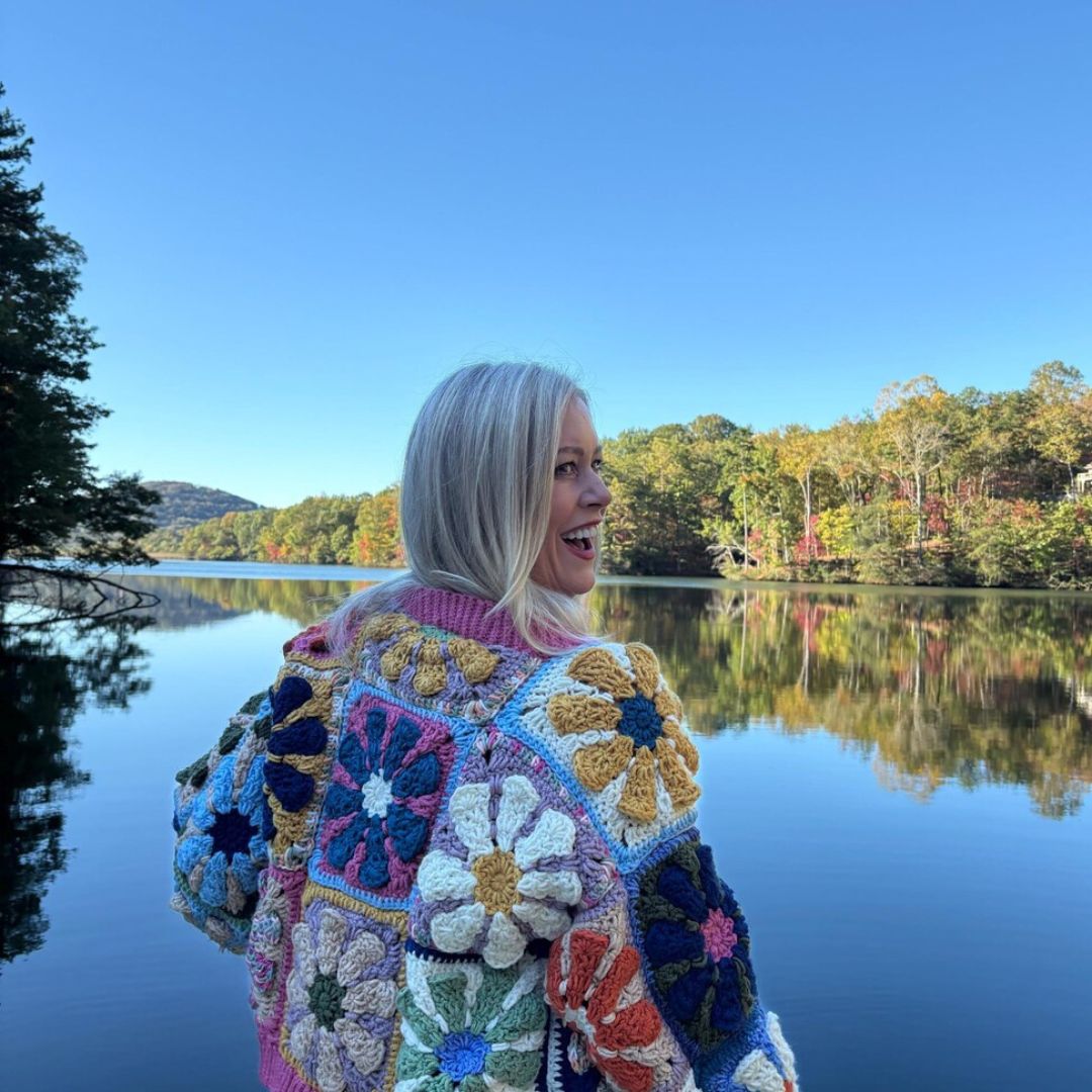 Person wearing a colorful retro daisy granny square crochet sweater by a lake with trees in the background