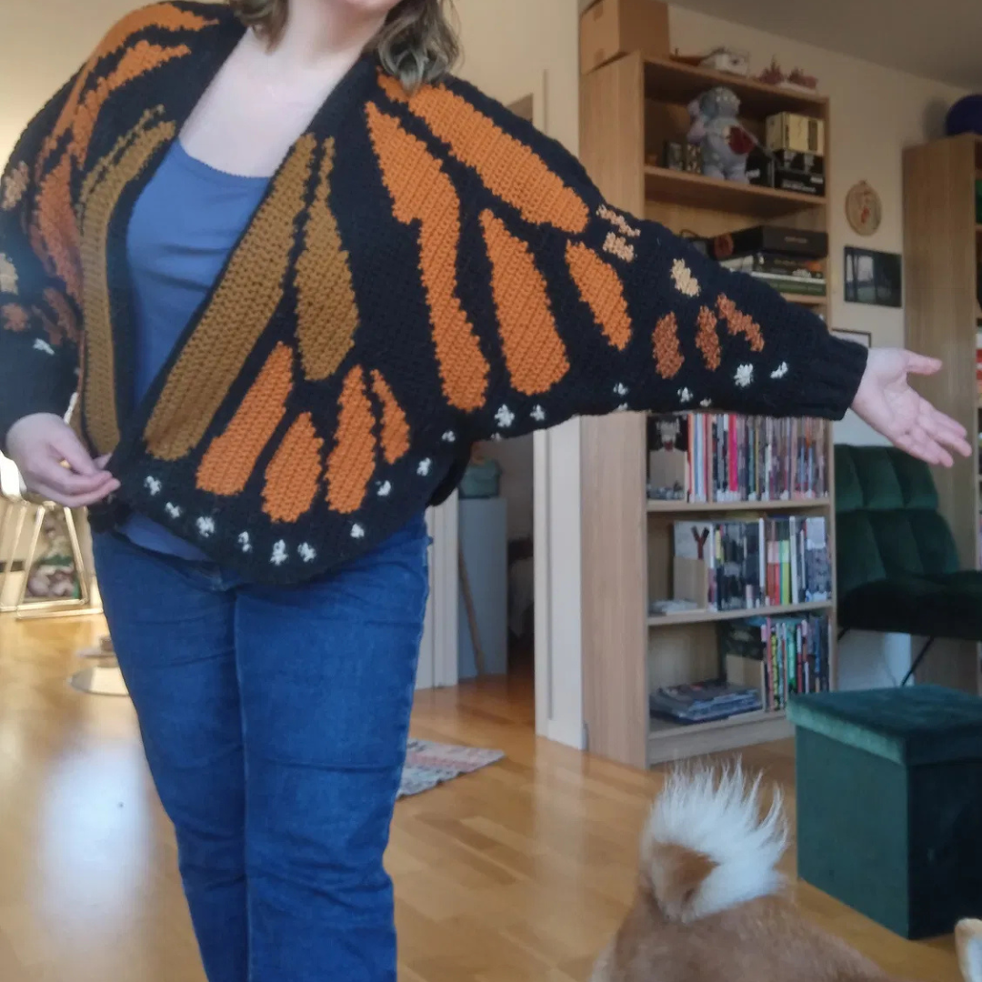 Person wearing a black and orange monarch butterfly patterned cardigan in a room with books and a dog.
